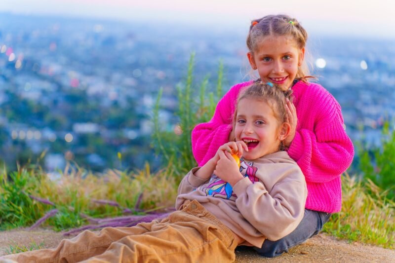 Two young sisters sitting together on a grassy hill overlooking a city panorama view at dusk, laughing and smiling.