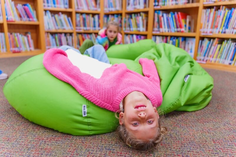 A playful young girl wearing a pink knit sweater lying upside down on a large green beanbag chair inside a library with bookshelves in the background.