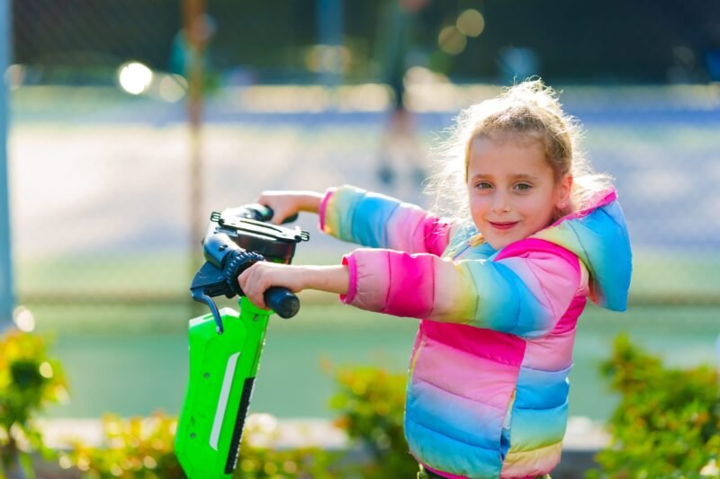 Close-up shot of a young girl smiling while riding a green and white electric scooter outside on a bright sunny day.