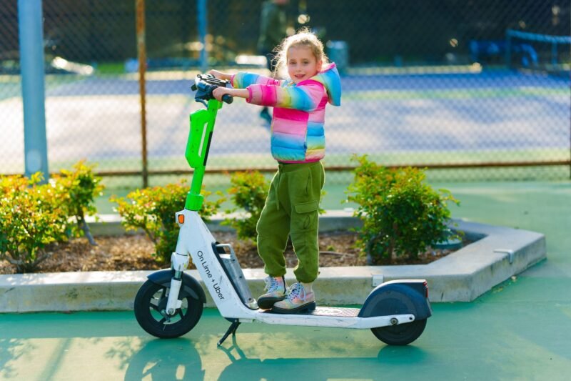 Full-length side view of a young girl in a colorful rainbow puffer jacket and green pants riding a white electric rental scooter outdoors.