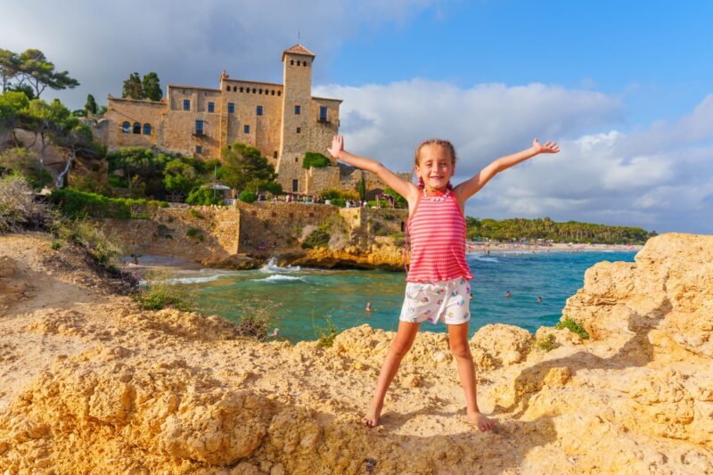 A happy young girl standing on a rocky sea shore with her arms wide open, featuring a historic stone castle and blue sea in Spain in the background.