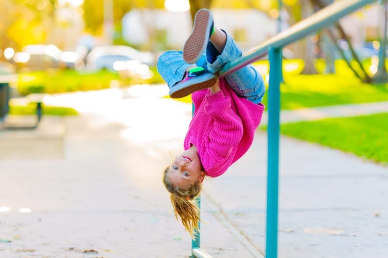 An energetic young girl hanging upside down from a metal playground handrail bar in a park on a sunny day.
