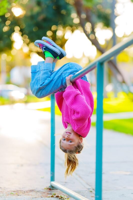 A happy young girl hanging upside down from a blue metal railing at a park, wearing a bright pink knit sweater and blue jeans.