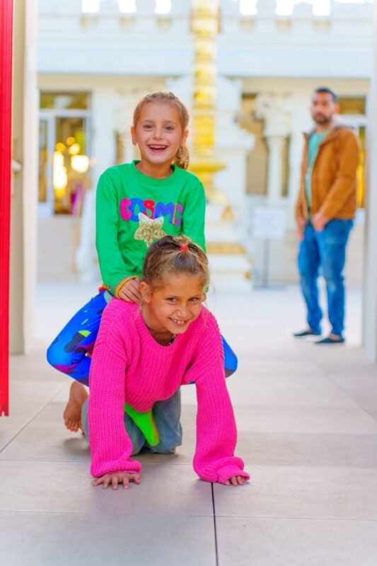 A cheerful girl giving her sister a piggyback ride on a tiled floor indoors, both smiling and wearing bright, colorful sweaters.