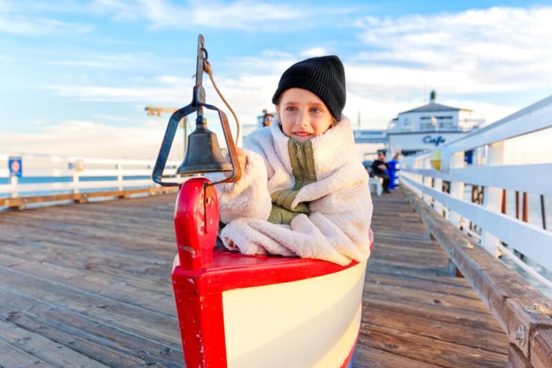 A young girl in a black beanie and oversized fleece jacket smiling while sitting in a decorative boat on a wooden pier next to a ship's bell.