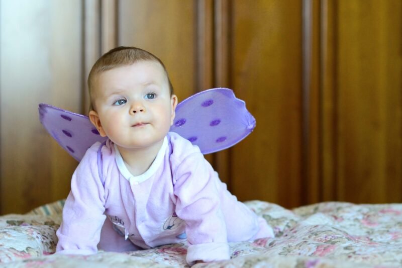 Baby wearing a light purple outfit and decorative purple butterfly wings crawling on a bed.