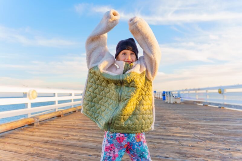A young girl playfully lifting the sleeves of a very large, oversized green and white fleece jacket while standing on a wooden pier.