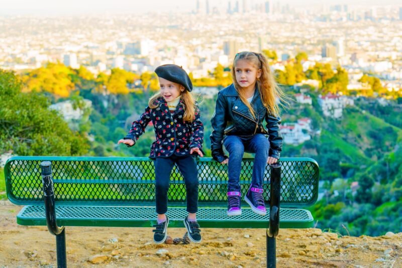 Two stylish young girls sitting on a green metal bench overlooking a scenic cityscape view at sunset.