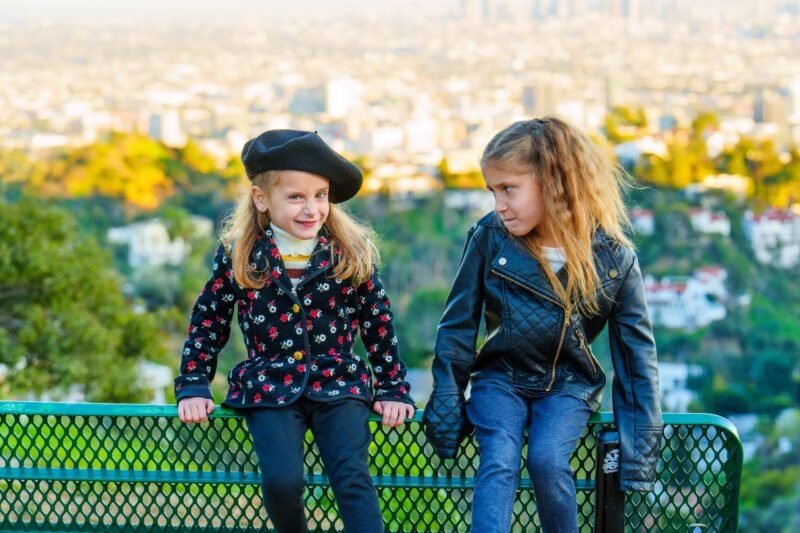 Two young girls sitting on a green park bench with a city view backdrop, one wearing a black beret and floral coat, the other in a black leather jacket.