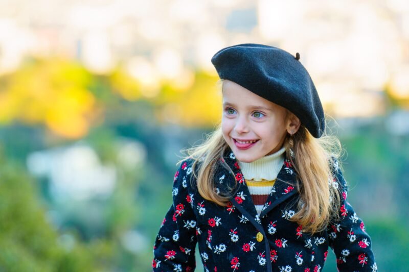 A close-up portrait of a smiling young girl wearing a classic black beret and a black coat with red and white floral patterns.