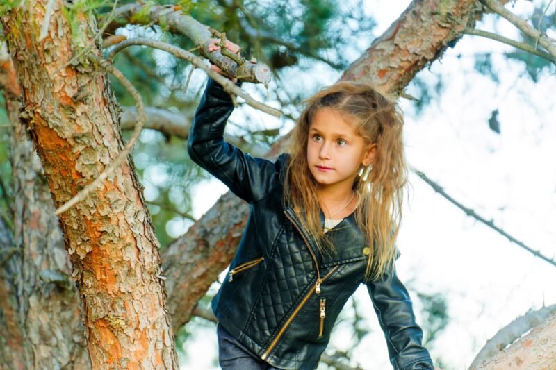 A young girl with long wavy hair wearing a black quilted leather jacket climbing a large pine tree.