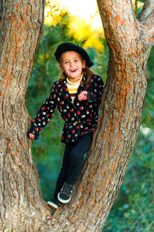 A laughing young girl in a black beret and floral jacket posing happily between the branches of a large pine tree.