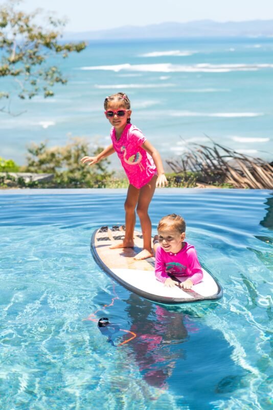 An older girl balances standing on a surfboard while a younger child lies on the front in a scenic infinity pool facing the ocean.