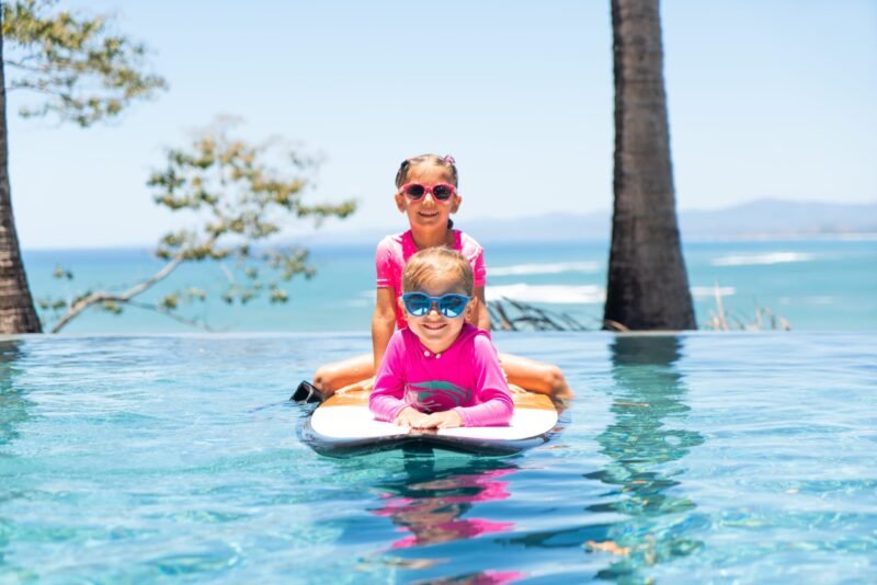 Two young girls wearing pink swimwear and sunglasses smile while playing together on a surfboard in a swimming pool next to the sea.