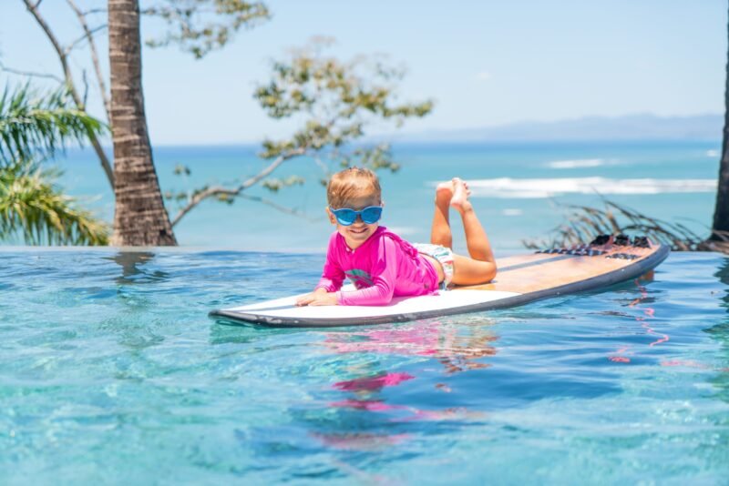 A smiling young child lies on their stomach with feet up on a surfboard inside a pool overlooking a sunny beach landscape.