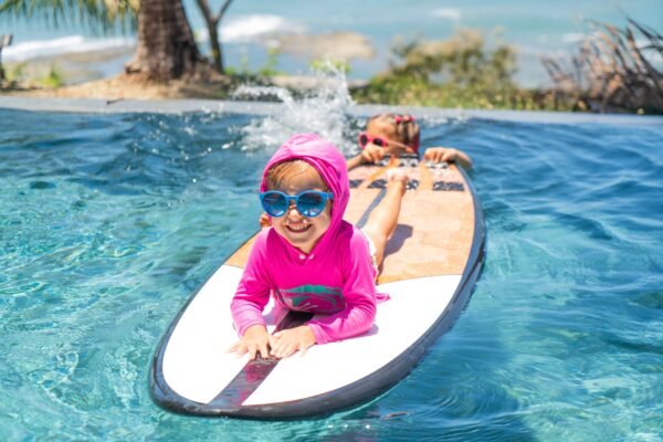 Close-up of a happy toddler wearing blue sunglasses and a pink hood lying on a surfboard in a pool, splashing water with a friend behind.