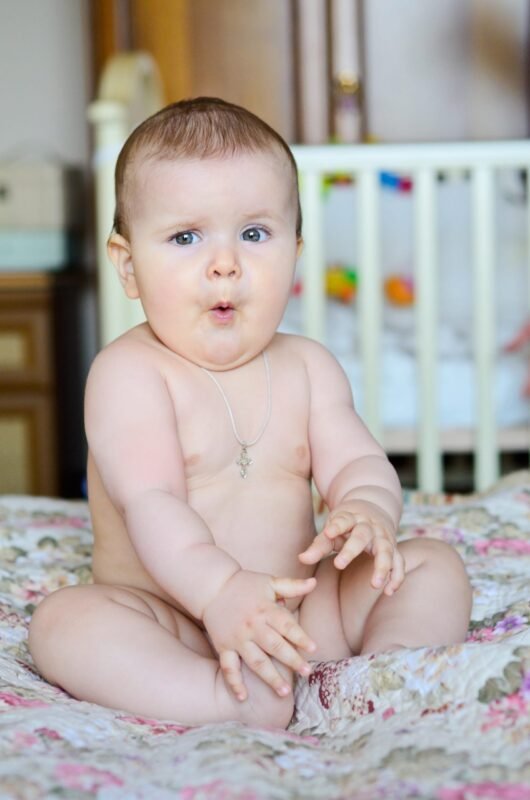 Chubby baby sitting on a floral blanket with a surprised facial expression, making a funny mouth shape