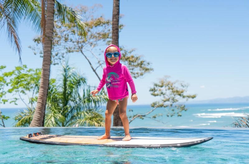 A young child wearing blue sunglasses and a pink hooded rash guard stands balanced on a surfboard inside an infinity pool overlooking the ocean.