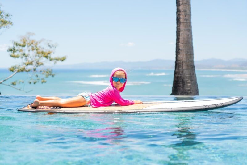 A toddler wearing a pink hooded sun shirt and blue sunglasses relaxes on a surfboard floating in a pool with an ocean view.