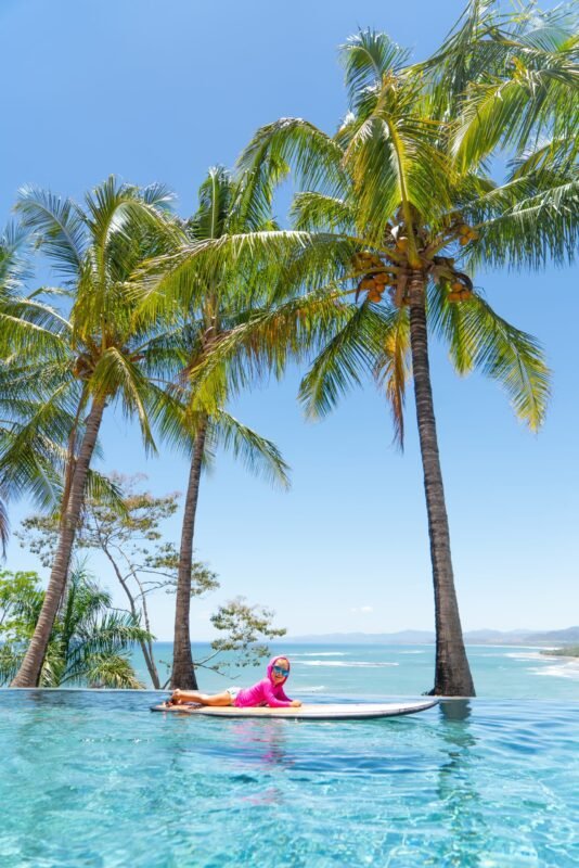 A young child in a pink swim shirt lies on a surfboard floating in a clear blue swimming pool with tall palm trees and an ocean beach in the background.