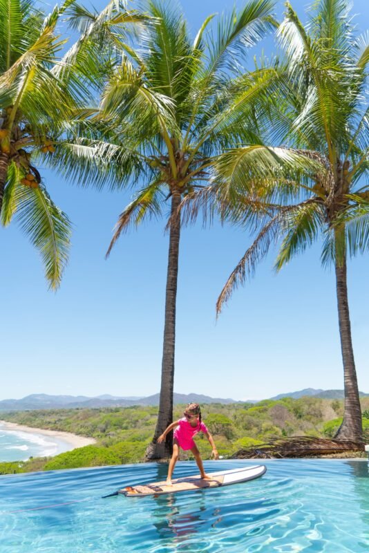 A young girl balances on a surfboard in a tropical infinity pool surrounded by tall coconut palm trees with a scenic coastline view.