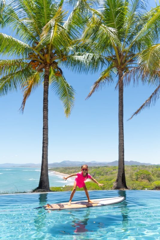 A young girl in bright pink swimwear stands with arms outstretched balancing on a surfboard in an infinity pool by the sea.