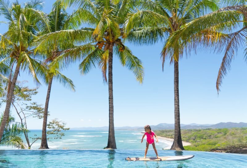Wide shot of a girl surfing on a board inside a luxury swimming pool with a panoramic view of palm trees and a sandy beach cove.
