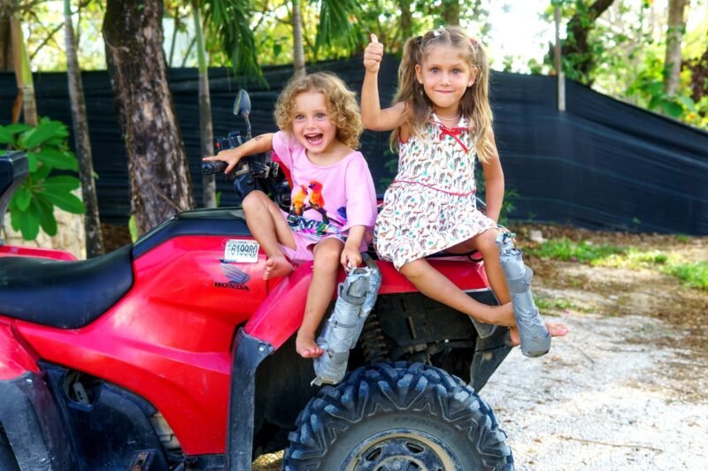 Two cheerful young girls sitting on a red ATV outdoors, one giving a thumbs up