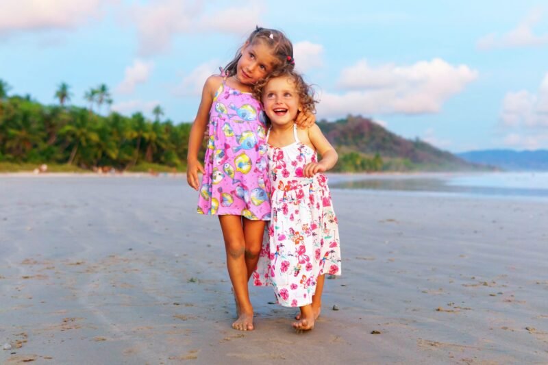 Two happy little girls in summer dresses walking together on a sandy beach with ocean background