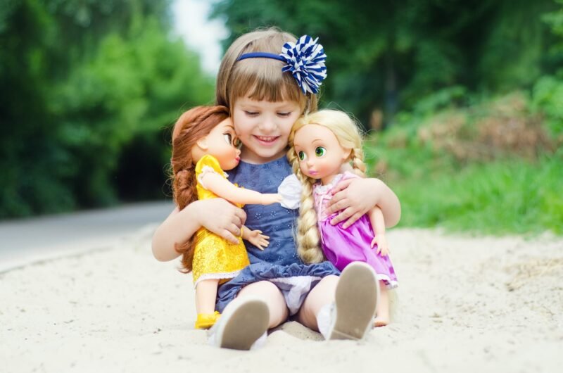 Smiling little girl sitting in a sandbox and hugging two dolls