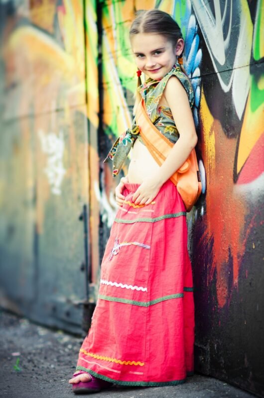 Young girl smiling while leaning against a colorful graffiti wall, wearing a long pink skirt and a green patterned top.