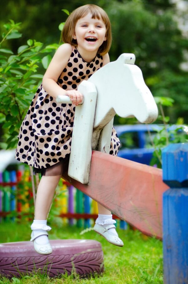 Laughing little girl in a polka dot dress riding a wooden horse on a playground