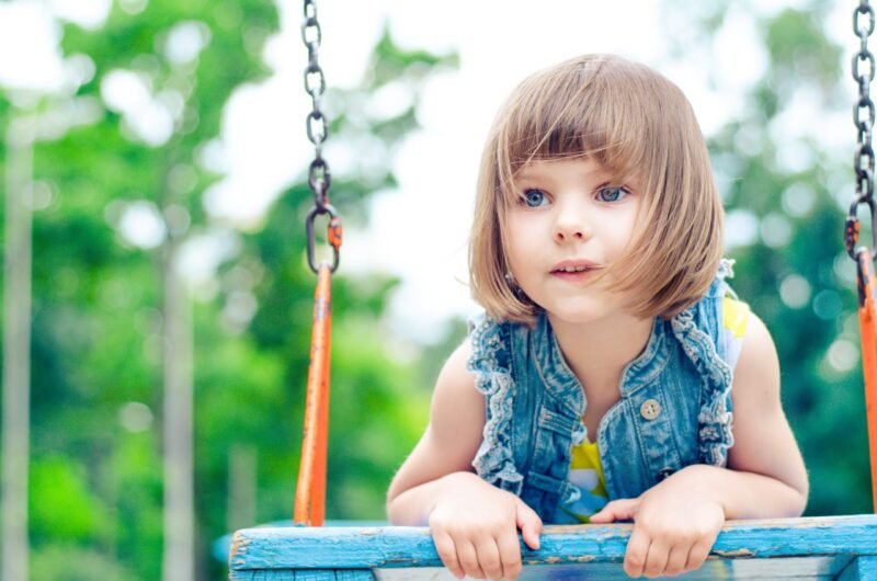 Little girl with a bob haircut leaning on a blue wooden swing at an outdoor playground