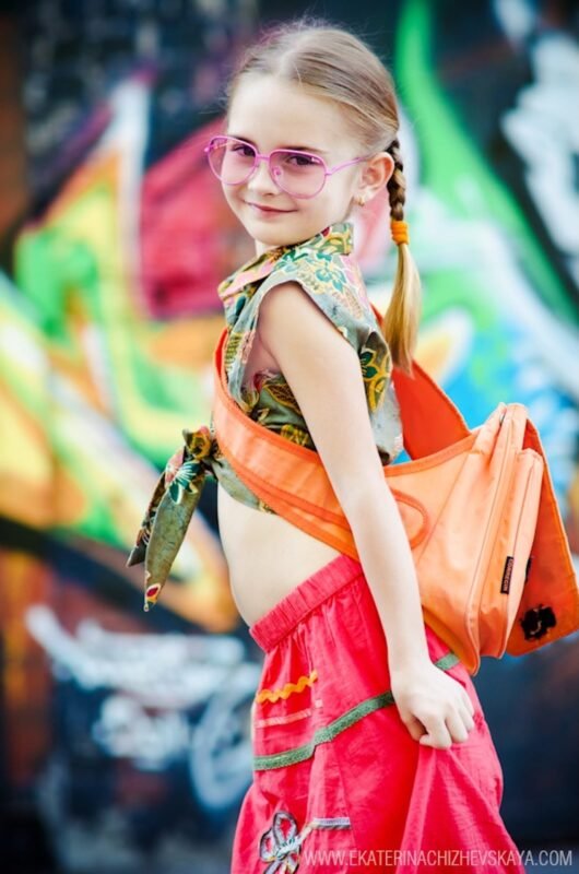 Young girl with braided hair and pink glasses posing in front of a colorful graffiti wall