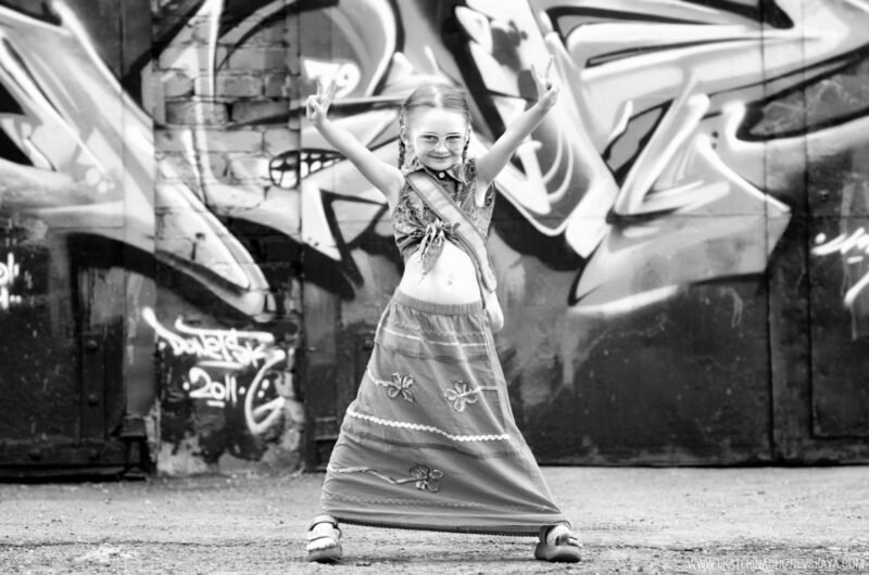 Black and white photo of a young girl in a long skirt posing with hands raised in front of graffiti
