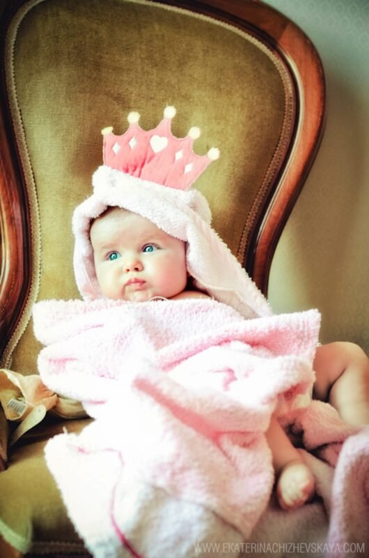 Adorable baby with blue eyes wrapped in a pink towel, wearing a toy princess crown while sitting on a vintage chair