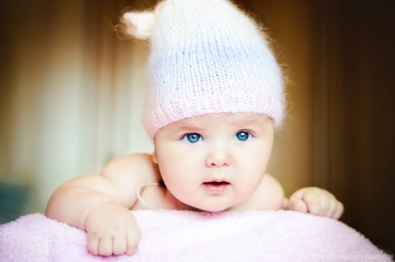 Close-up of a cute baby with bright blue eyes lying on their stomach wearing a soft knitted winter hat