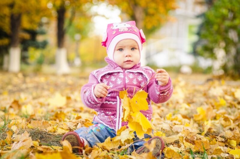 Cute toddler in a pink jacket and hat sitting outside on the ground covered with yellow autumn leaves