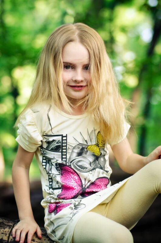 Young blonde girl sitting on a fallen tree log in a green forest, wearing a yellow t-shirt and leggings