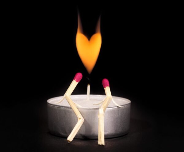 Conceptual match art showing two bent matchsticks resembling a couple sitting together on a tealight candle with a heart-shaped flame above them