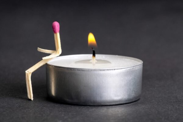 A creative matchstick figure shaped like a sitting person leaning against a burning white tea light candle.