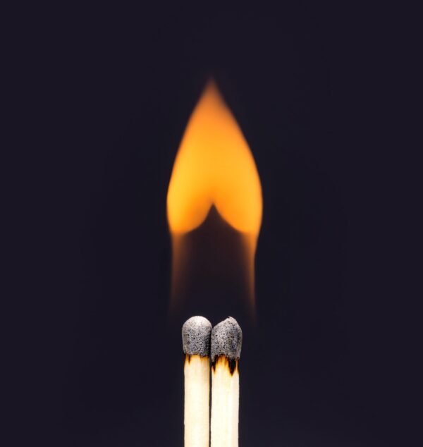 Two matchsticks standing close together with their flames merging into a single heart-shaped flame against a dark background