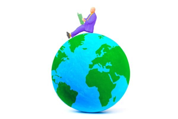 Miniature businessman figure sitting on top of a globe model and reading a book against a white background.