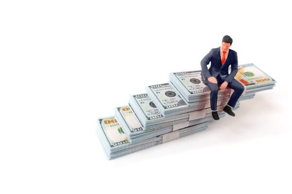 Miniature figure of a businessman in a suit sitting on a staircase made of 100 dollar bill stacks.