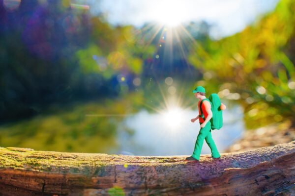 Miniature hiker figurine walking along a mossy log with a sunlit river or lake and green trees blurred in the background