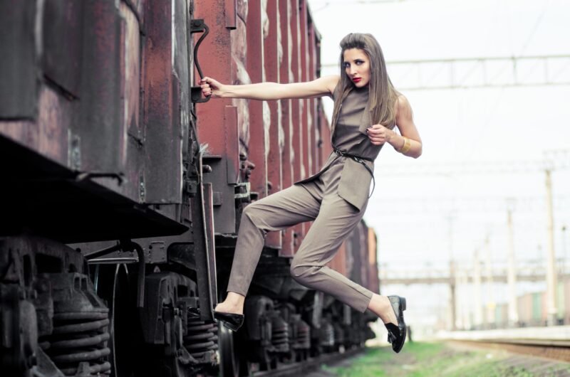 Fashion model in a khaki jumpsuit jumping and posing dramatically on the side of a vintage train car at a railway station.