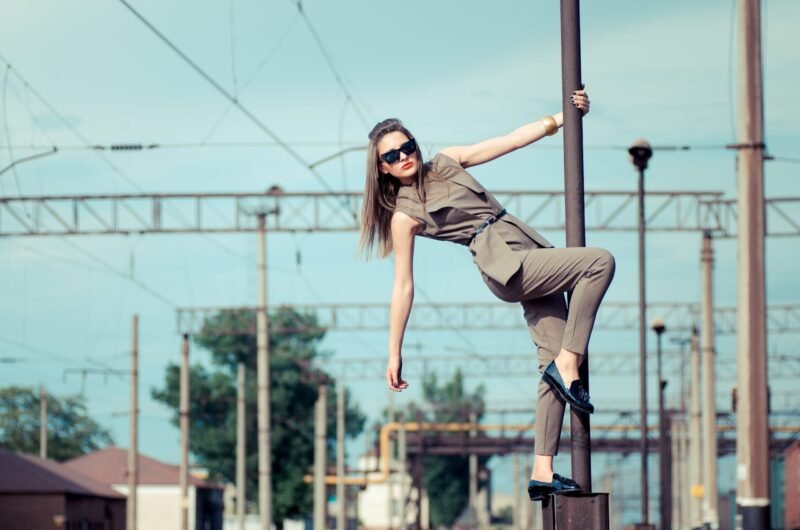 Stylish woman wearing sunglasses and a beige jumpsuit posing dynamically on a metal pole at a railway station