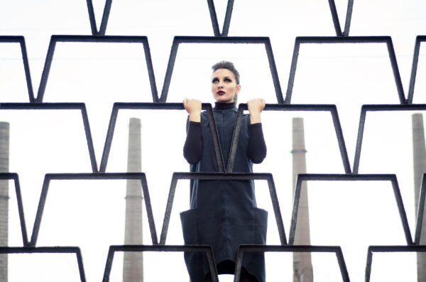 Avant-garde fashion portrait of a woman looking through a geometric black metal fence with industrial chimneys in the background