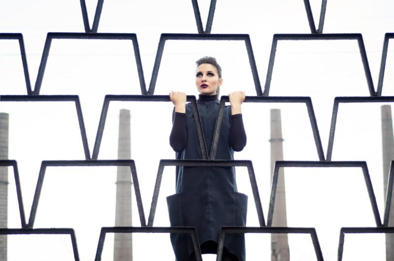 Avant-garde fashion portrait of a woman looking through a geometric black metal fence with industrial chimneys in the background