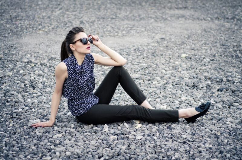 Side view of a fashionable woman in a patterned sleeveless top and sunglasses sitting on a gravel surface